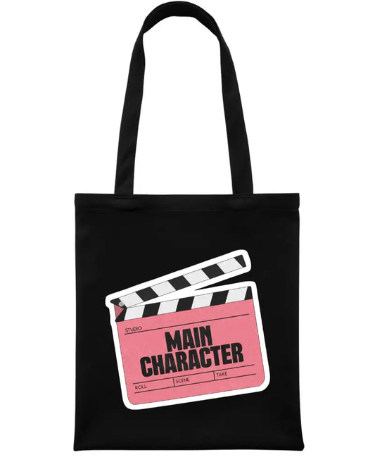 A black canvas tote bag with two black shoulder straps, featuring a graphic print of a film clapperboard with a pink 'MAIN CHARACTER' label on the front.