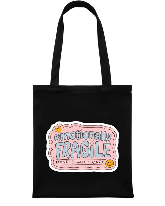 A black canvas tote bag with two shoulder straps, featuring a graphic label on the front that reads 'emotionally FRAGILE HANDLE WITH CARE' in pastel pink, blue, yellow, and orange tones.