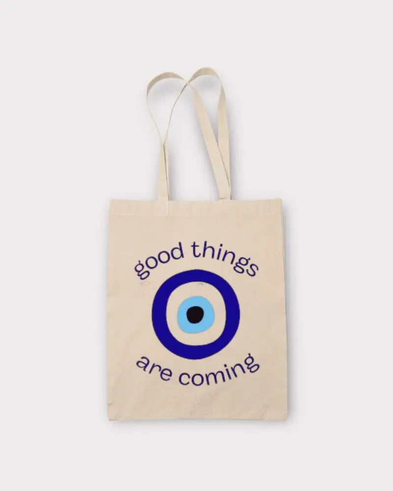 A natural canvas tote bag with two shoulder straps, featuring a blue and white evil eye graphic and the text 'good things are coming' in dark blue, set against a light gray background.