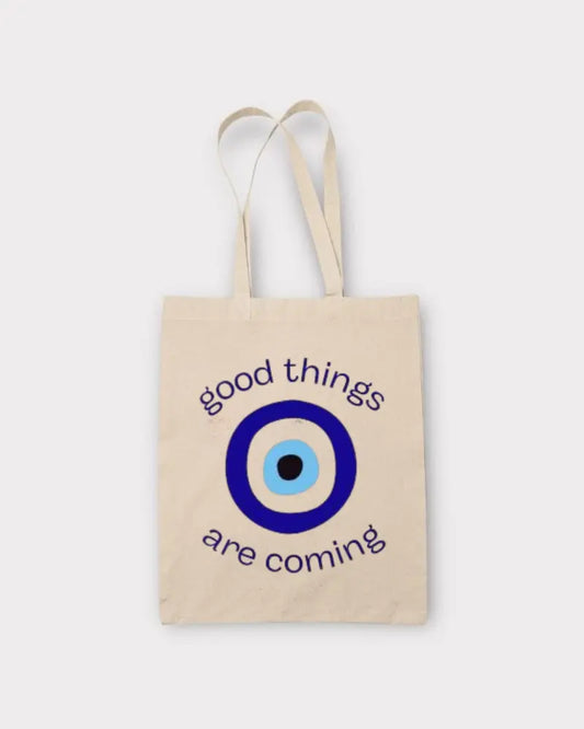 A natural canvas tote bag with two shoulder straps, featuring a blue and white evil eye graphic and the text 'good things are coming' in dark blue, set against a light gray background.